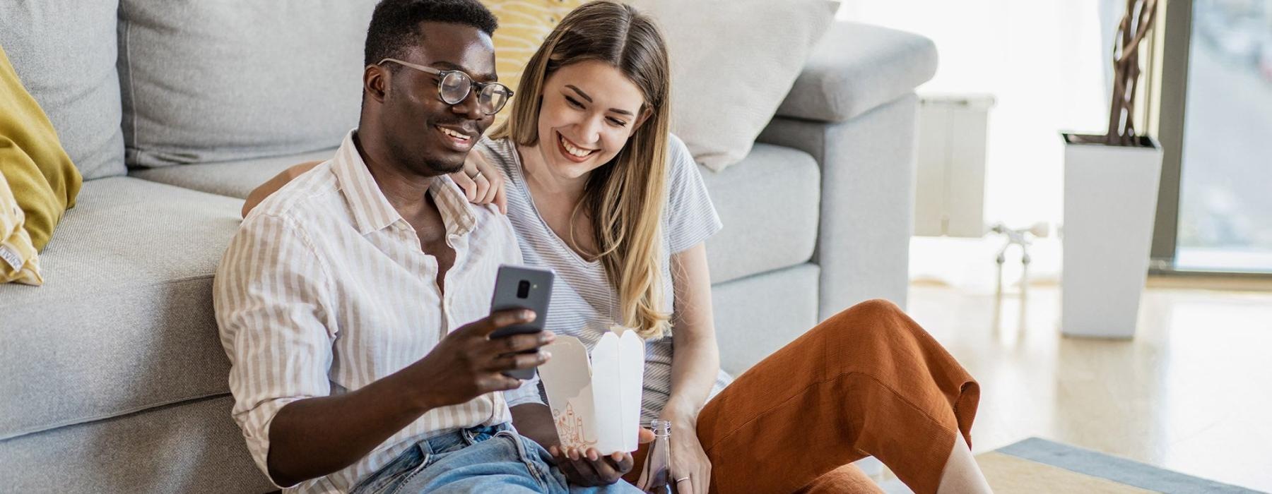 a man and woman with take out, sit against a couch on their living room floor and watch at their cell phone